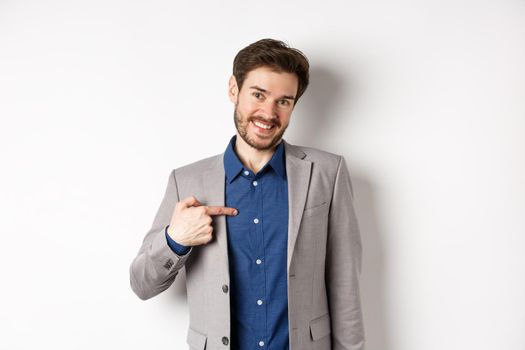 Modest smiling man in suit pointing at himself with cute face, self-promoting, standing on white background.