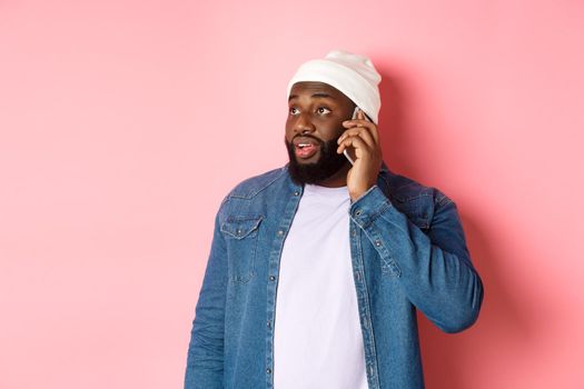 Hipster Black man talking on phone, looking left and having mobile conversation, standing over pink background.