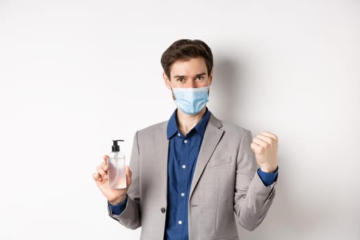 Covid-19, pandemic and business concept. Cheerful office guy in medical mask and suit, looking motivated, showing bottle of antiseptic, white background.