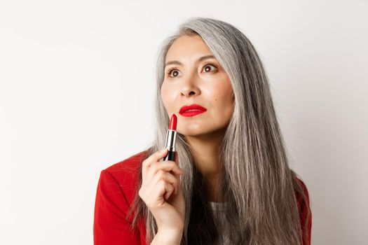 Beauty and makeup concept. Sensual mature asian female with grey hair, looking aside and showing red lipstick, standing over white background.
