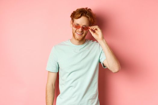 Tourism and vacation concept. Relaxed young man with red hair, standing in sunglasses and t-shirt and smiling pleased, standing over pink background.