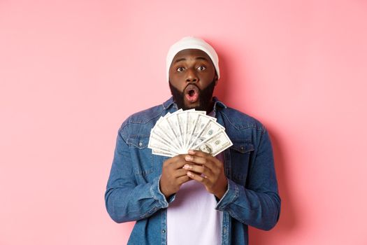 Amazed african-american man receive cash prize, showing money and staring at camera in awe, standing over pink background.