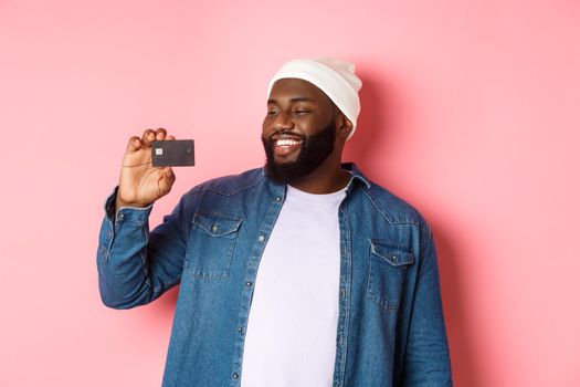 Shopping concept. Image of happy african-american man looking satisfied at credit card, recommending bank, standing over pink background.