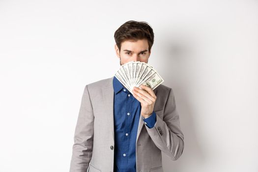 Rich businessman in suit showing dollar bills, hiding face behind money with sensual gaze at camera, white background.