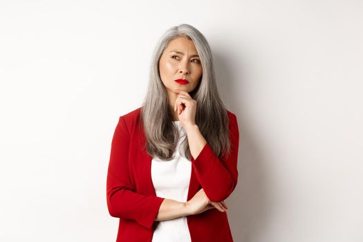 Business concept. Thoughtful senior asian businesswoman looking aside, thinking while standing over white background.