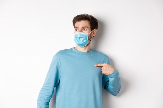 Covid-19, health and quarantine concept. Confused man in medical mask pointing at himself, looking left with ambushed face, standing over white background.