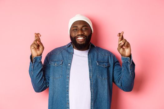 Optimistic african-american man making wish, holding fingers crossed and smiling, standing over pink background.