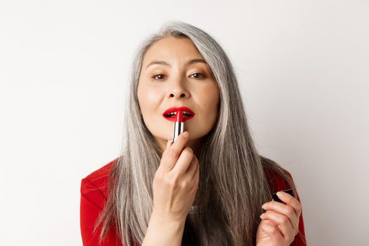 Beauty and fashion concept. Close up of senior asian woman looking in mirror and apply red lipstick, standing over white background.