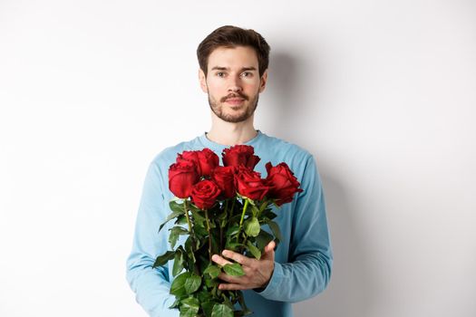 Confident young man bring flowers on Valentines day date, holding romantic bouquet, standing over white background.