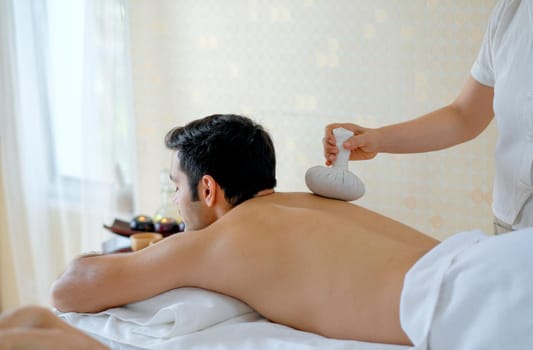 Handsome man enjoying with massage by herbal ball on his back in spa room with day light.