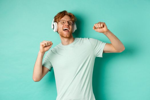 Lifestyle concept. Carefree guy with red hair, dancing and having fun, listening music in headphones, standing over turquoise background.