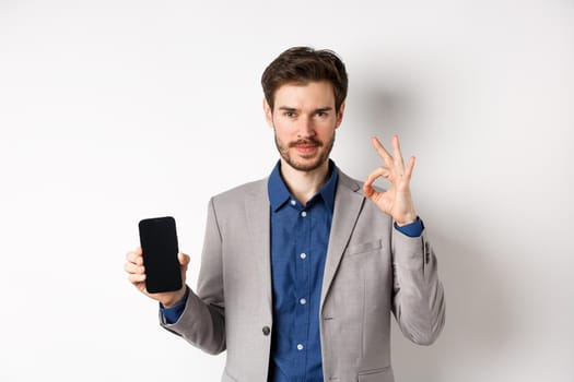 E-commerce and online shopping concept. Confident handsome business man in suit showing Ok sign and empty smartphone screen, recommend application, white background.