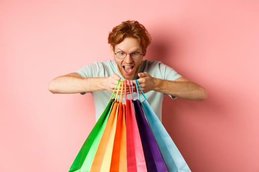 Excited young man, shopper holding shopping bags and smiling happy, looking excited at purchased items, standing over pink background.