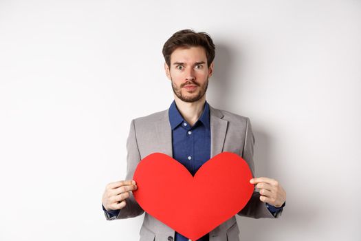 Excited caucasian man in suit looking at camera, holding big red heart cutout on valentines day, standing over white background. Copy space