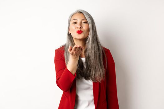 Elegant asian woman with red blazer and lips, blowing air kiss at camera, concept of valentines day and romance, white background.
