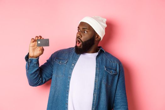 Shopping concept. Image of bearded african-american man staring shocked at credit card, standing against pink background.
