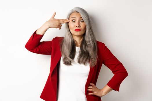 Annoyed and sarcastic asian female entrepreneur shooting herself with finger gun, staring bothered at camera, standing against white background.