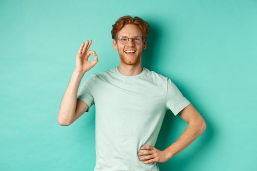 Happy young man with red hair and beard, wearing glasses and t-shirt, smiling satisfied and showing ok sign, say yes, approve and agree, standing over turquoise background.