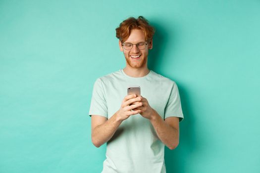 Handsome young man in glasses with red messy hair reading message on mobile phone, smiling and looking at screen, standing over mint background.