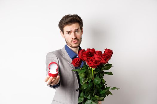 Handsome boyfriend in suit asking to marry him, standing with red bouquet of roses and engagement ring, looking pleading at camera, standing over white background.