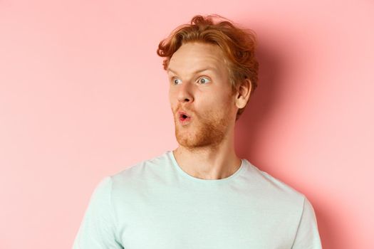 Headshot portrait of surprised redhead man with beard, looking left and saying wow, raising eyebrows amazed, standing over pink background.