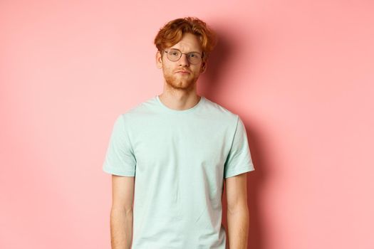 Sad and gloomy redhead bearded man in t-shirt and glasses, staring at camera bored and unamused, standing over pink background.