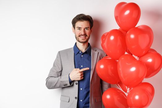 Lovers day. Handsome smiling guy in suit making surprise gift on Valentines date, pointing at romantic heart balloons and looking happy, standing over white background.