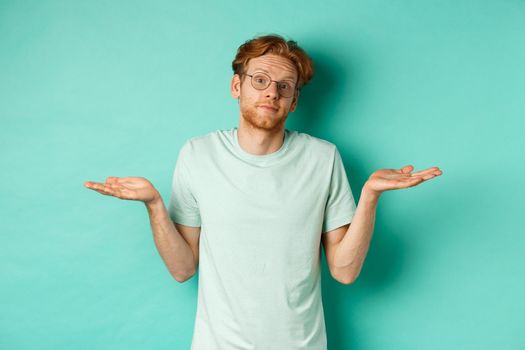 Portrait of confused redhead man in t-shirt and glasses know nothing, shrugging shoulders and looking clueless at camera, standing against turquoise background.