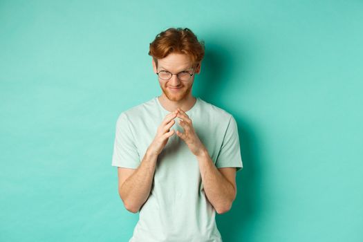 Devious redhead man in glasses and t-shirt pitching an idea, steeple fingers and look from under forehead with sly and smug smile, standing over mint background.