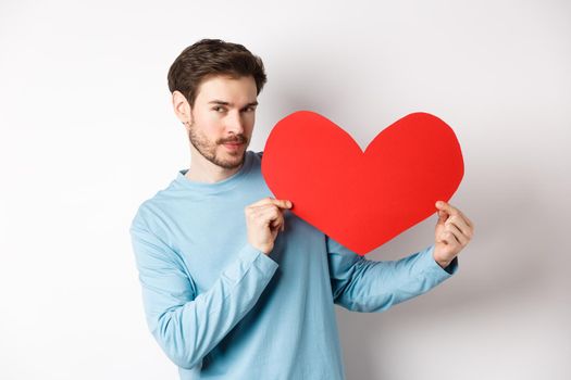 Valentines day. Handsome and romantic man holding big red valentine heart cutout, looking seductive at camera, making love confession, white background.