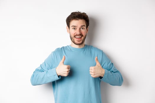 Excited smiling man showing thumbs up to praise excellent product, looking amazed and happy at camera, recommending good deal, standing on white background.