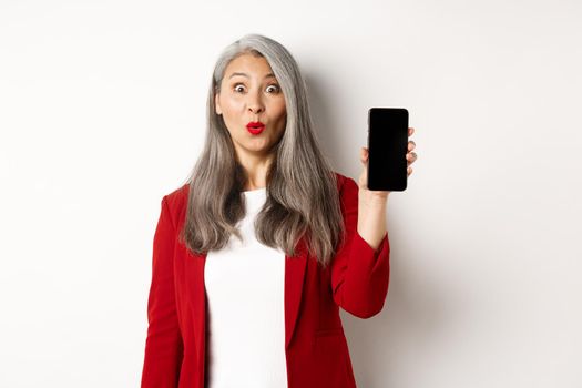 Amused senior woman in red blazer, looking fascinated and showing blank smartphone screen, standing over white background.
