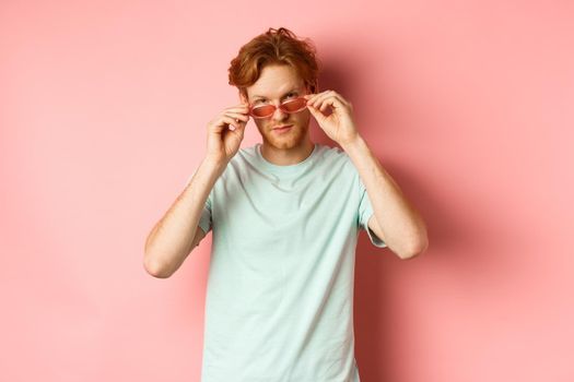 Handsome redhead man in summer sunglasses looking sassy at camera, standing over pink background.