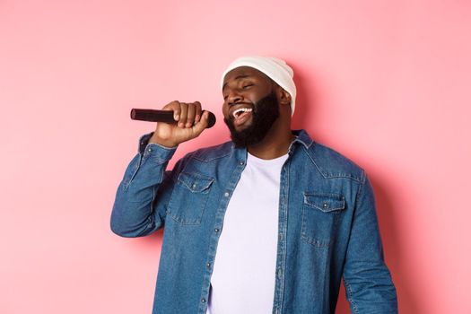 Handsome Black man in beanie and denim shirt singing karaoke, holding microphone, standing over pink background.
