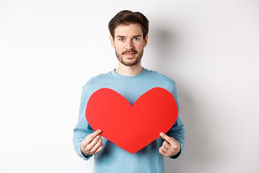 Smiling young man holding valentines heart cutout and looking at camera, waiting for true love girlfriend, standing over white background.