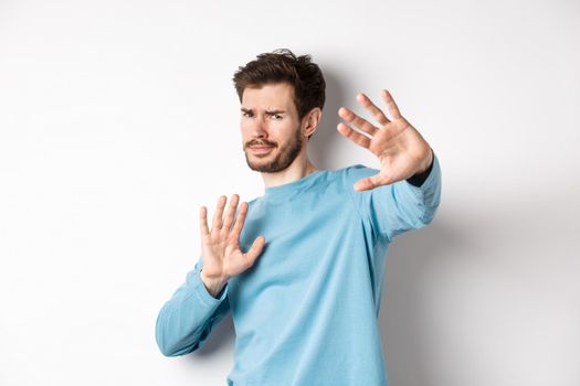 Stay away from me. Reluctant young man step back with hands stretch out in defensive gesture, protecting himself, standing over white background.