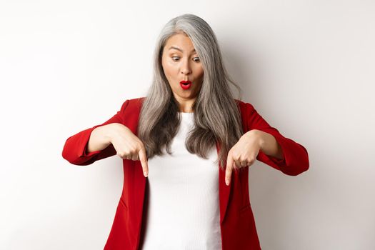 Impressed asian mature woman checking out promo, pointing and looking down with amazed face, standing in blazer over white background.