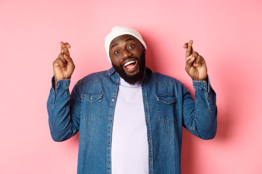 Optimistic african-american man making wish, holding fingers crossed and smiling, standing over pink background.