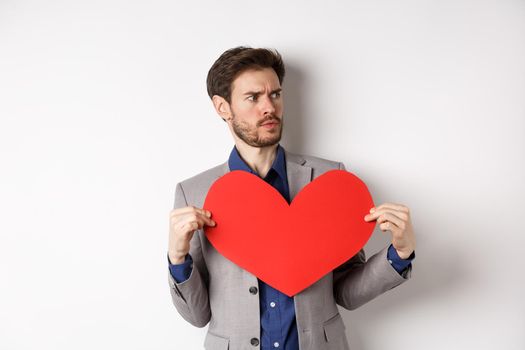 Confused man in suit holding big red heart and looking right, searching for love on Valentines day, standing over white background.