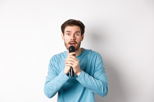 Confused young man looking nervously at camera while singing karaoke, holding microphone, standing over white background.