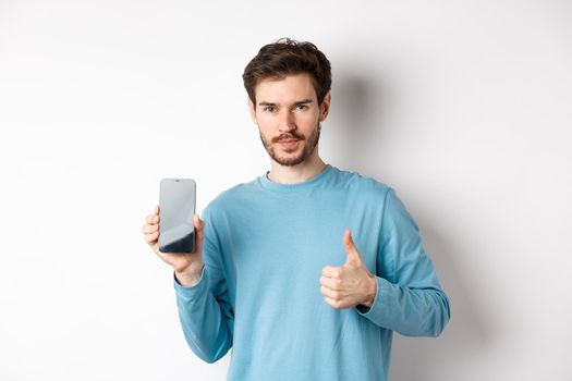 Young man showing empty smartphone screen and thumb up, recommend mobile application or online shopping offer, standing over white background.