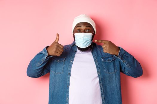 Coronavirus, lifestyle and global pandemic concept. Young african-american man pointing at face mask and showing thumbs-up, protect himself from covid, pink background.