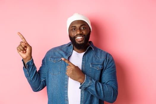 Happy african american man pointing fingers upper left corner, showing promo offer logo, smiling pleased, wearing beanie with denim jacket, pink background.
