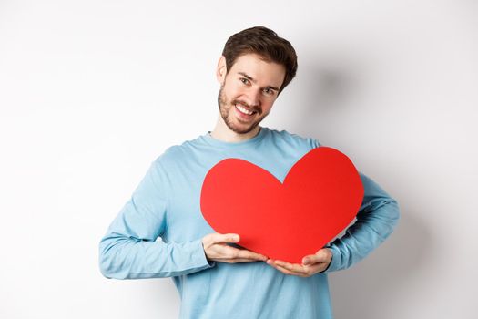 Charming lover man holding Valentines day red heart and smiling, I love you gesture, standing over white background.