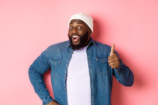 Happy african-american male model showing thumb-up and saying yesm smiling satisfied, standing over pink background.