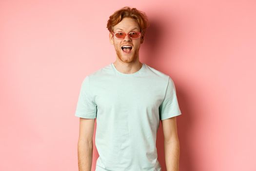 Image of surprised redhead man on vacation, wearing sunglasses with summer t-shirt, open mouth and saying wow amazed, standing over pink background.