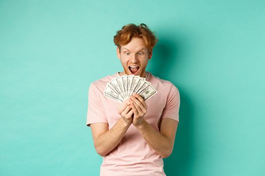 Excited young man winning prize money, counting cash and looking amazed at dollars, standing over turquoise background.