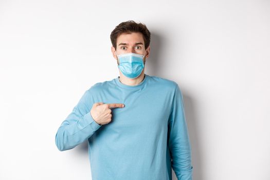 Coronavirus, health and quarantine concept. Surprised guy in medical mask points at himself, standing over white background.