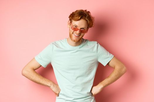 Tourism and vacation concept. Funny bearded man with red hair, wearing glasses, smiling and looking from under forehead with hands on hips, standing over pink background.