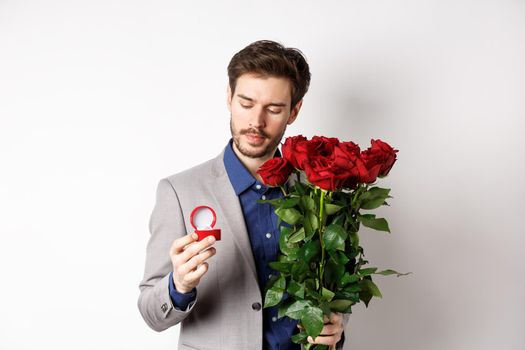 Romantic man in suit looking pensive at engagement ring, going to make a marriage proposal on Valentines day, holding bouquet of roses, standing over white background.